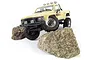 A creme-colored 1/18 scale HPI Venture18 RC Toyota Hilux SR5 rock crawler climbing over two large, textured rocks on a white background.