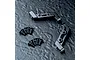 Two black anodized aluminium bulkhead parts for an RC car, accompanied by eight small black countersunk screws, arranged on a dark textured surface.