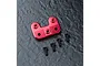 A red anodized aluminium steering arm mount for an RC car, accompanied by four black flat-head screws on a dark textured surface.
