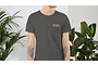A person wearing a dark grey T-shirt with a small TDK logo on the left chest, standing between two indoor potted plants.
