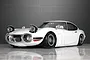 A front three-quarter view of a white Toyota 2000GT RC body shell with pop-up headlights open, parked on a reflective surface.