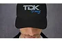 A person wearing a black dad hat featuring an embroidered TDK Racing logo on the front.