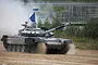 A Russian T-72B3M main battle tank driving through a dusty field with a blue flag and lights mounted on the turret.