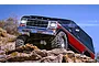 A Traxxas TRX-4 Ford Bronco RC crawler driving over rocky terrain with its bright LED headlights illuminated against a clear blue sky.