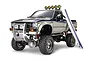 A 1/10 scale Tamiya Toyota Hilux Highlift RC truck with a surfboard leaning against the side, featuring a silver and black body and off-road tires.