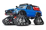 A blue Traxxas TRX-4 RC rock crawler truck equipped with Traxx all-terrain tracks instead of wheels, featuring a roof rack and red fuel canisters.