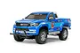 A studio shot of a blue Tamiya Toyota Hilux Extra Cab RC truck kit on a white background, featuring chrome wheels and detailed racing decals.