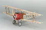 A partially assembled wooden model of a WWI Sopwith Camel biplane fighter, showing the internal rib structure of the wings and a red engine cowling.