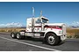 A white Western Star 4964 semi-truck model kit, featuring red star decals and chrome details, shown on a road against a desert landscape background.