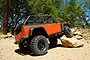 An orange Axial SCX10 Dingo RC rock crawler driving over rocks in a wooded outdoor area.