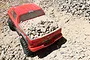 A red Axial SCX10 Ram Power Wagon RC rock crawler driving on a rocky dirt path with a bed full of small rocks.
