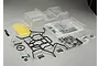 An unpainted clear polycarbonate RC crawler body shell kit with black plastic roll cage parts, light buckets, and decal sheets on a grey background.
