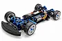 A Tamiya TB-08 Evo 1/10 scale RC touring car chassis featuring blue anodized aluminum parts, black carbon fiber components, and white wheels.