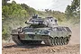 A Leopard 1 A5 tank in camouflage paint driving through a dusty, wooded landscape.
