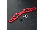 A red anodized aluminium bumper brace for an RC car, accompanied by two black countersunk screws and a small MST brand sticker on a dark textured surface.