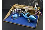 Italeri modelling workstation featuring a wooden organizer stand and a blue A3 cutting mat, holding various model paints, brushes, and a scale race car model.